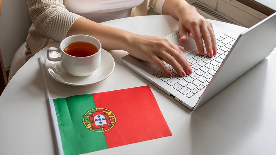 Lonely woman freelancer with flag of Portugal enjoying having breakfast with cup of coffee working on laptop sitting near window in cafe at morning.