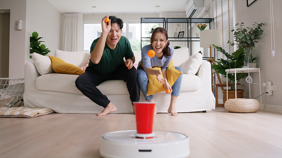 Internet of things smart urban house IOT appliances better life. Asia people man woman joy win fun beer ping pong ball throw cup game to robot hoover clean floor at home sofa living room with pet dog.